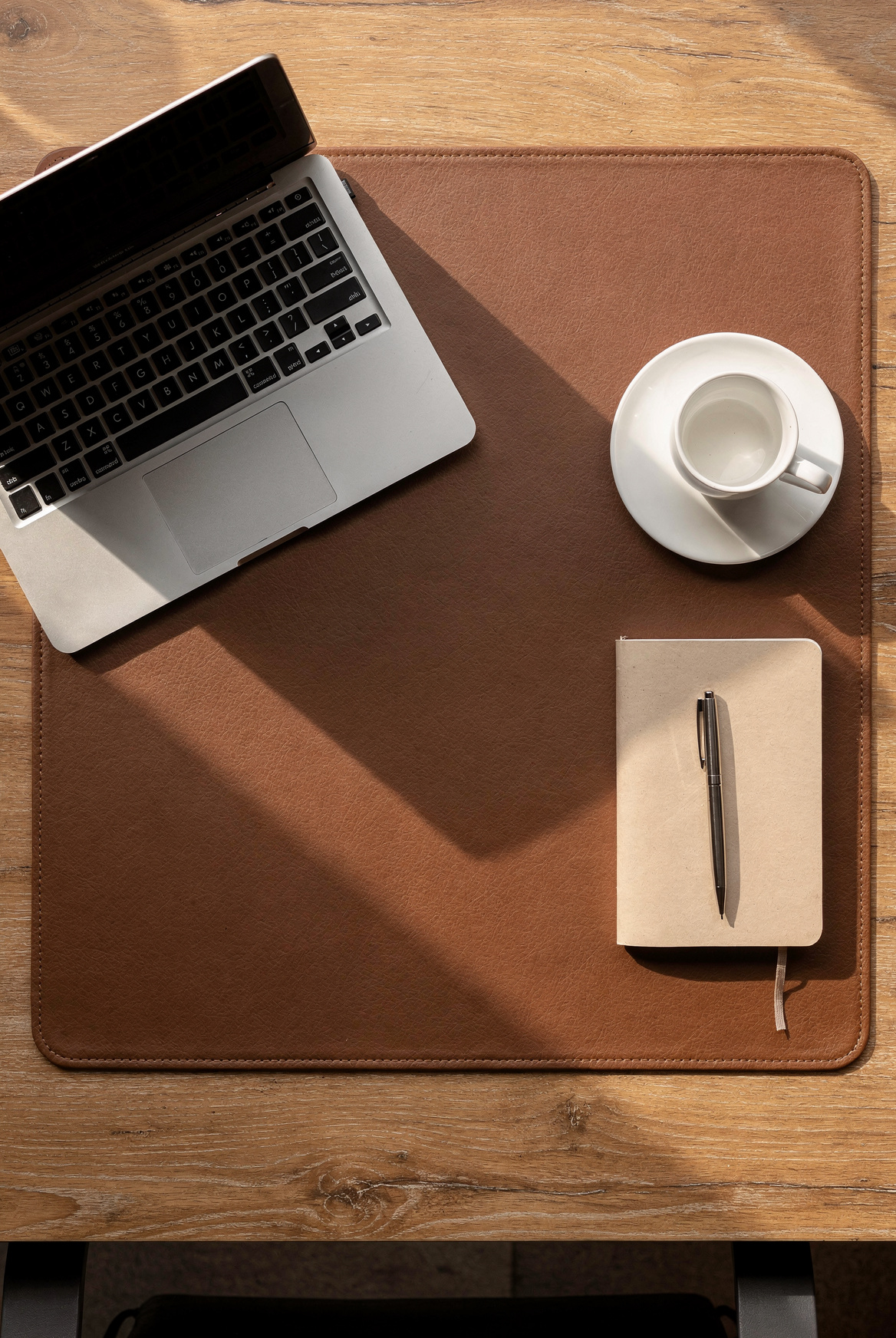 A large brown faux leather desk pad with stitched edges on a wooden desk, open laptop and ceramic coffee mug on it, soft morning daylight, warm beige tones, clean and organized, editorial home office style