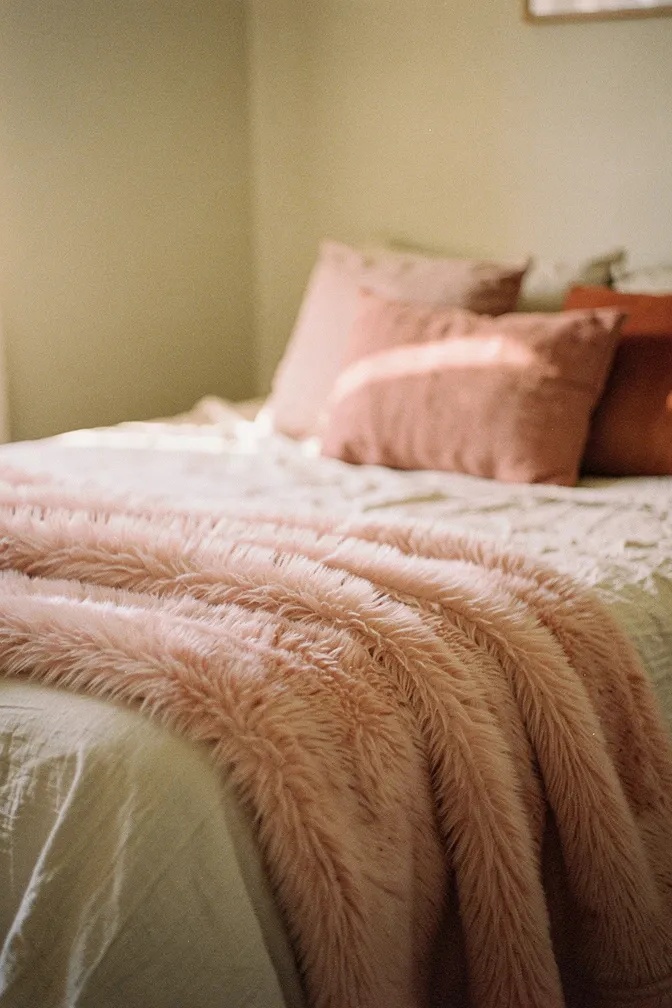 Light pink faux fur throw blanket draped at foot of bed, blush and terracotta pillows, cozy bedroom
