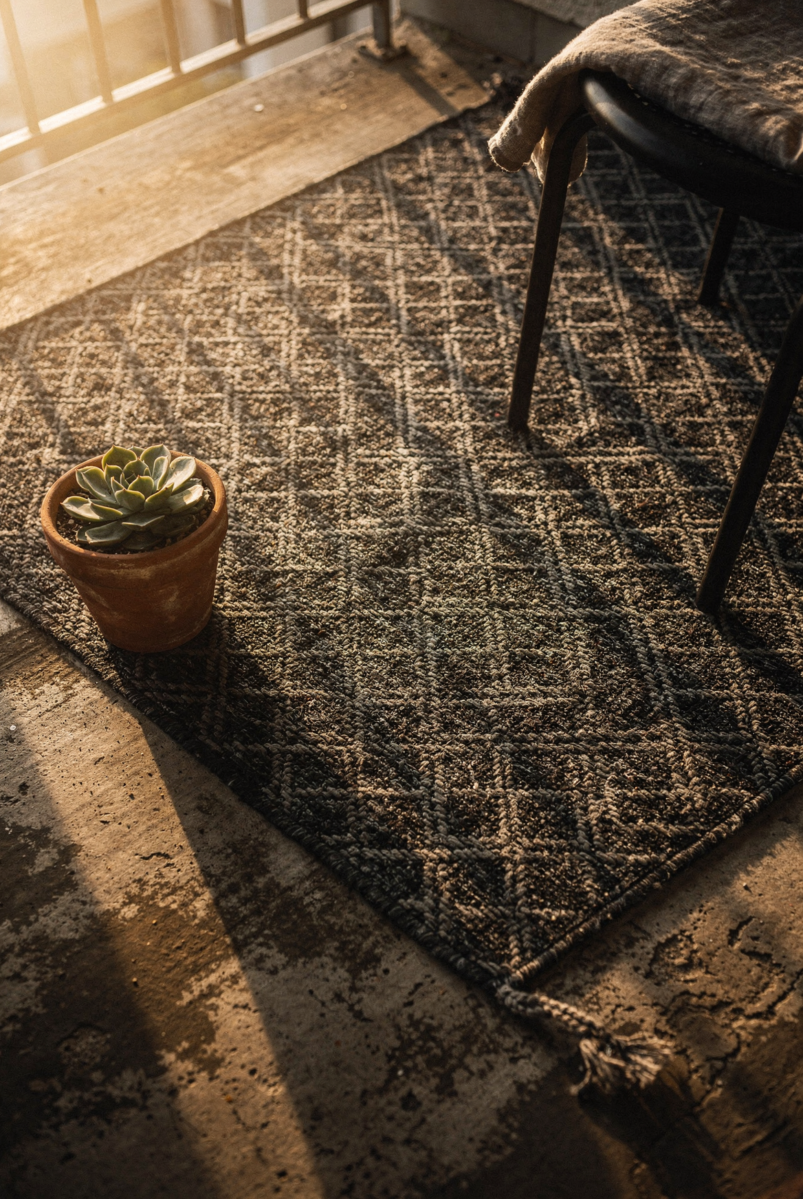 SAND MINE black and grey lattice outdoor rug defining an apartment balcony floor, bistro chair leg at edge, terracotta succulent pot at corner, morning light