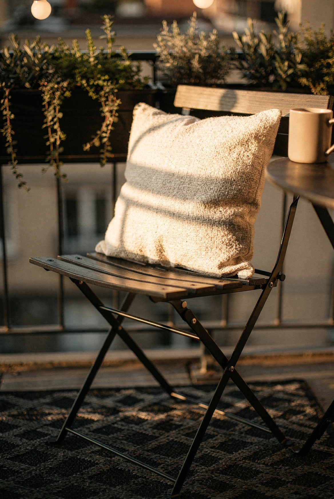 Metal bistro chair with MIULEE cream farmhouse waterproof pillow cover, SAND MINE lattice rug beneath, railing planters with herbs in background, warm afternoon golden light