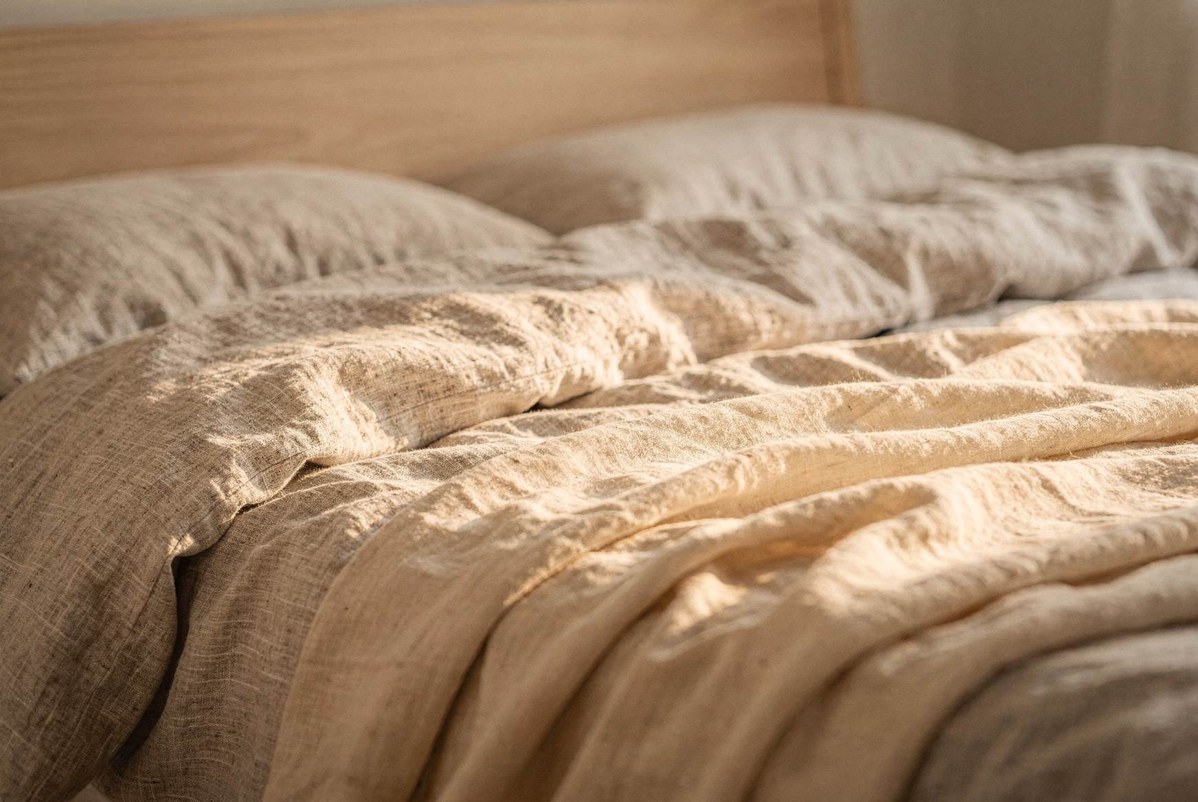 Stonewashed linen bedding in warm white on a simple wooden bed frame, natural light from sheer curtains, a folded quilt at the foot, relaxed morning aesthetic, editorial interior photography