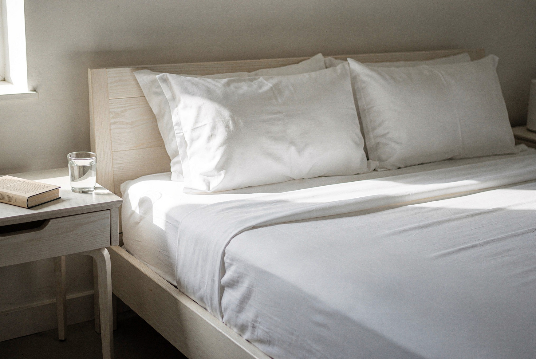 Crisp white cotton percale sheets on a hotel-style bed, neatly tucked, natural morning light, minimal bedside table with a book and water glass, editorial interior photography