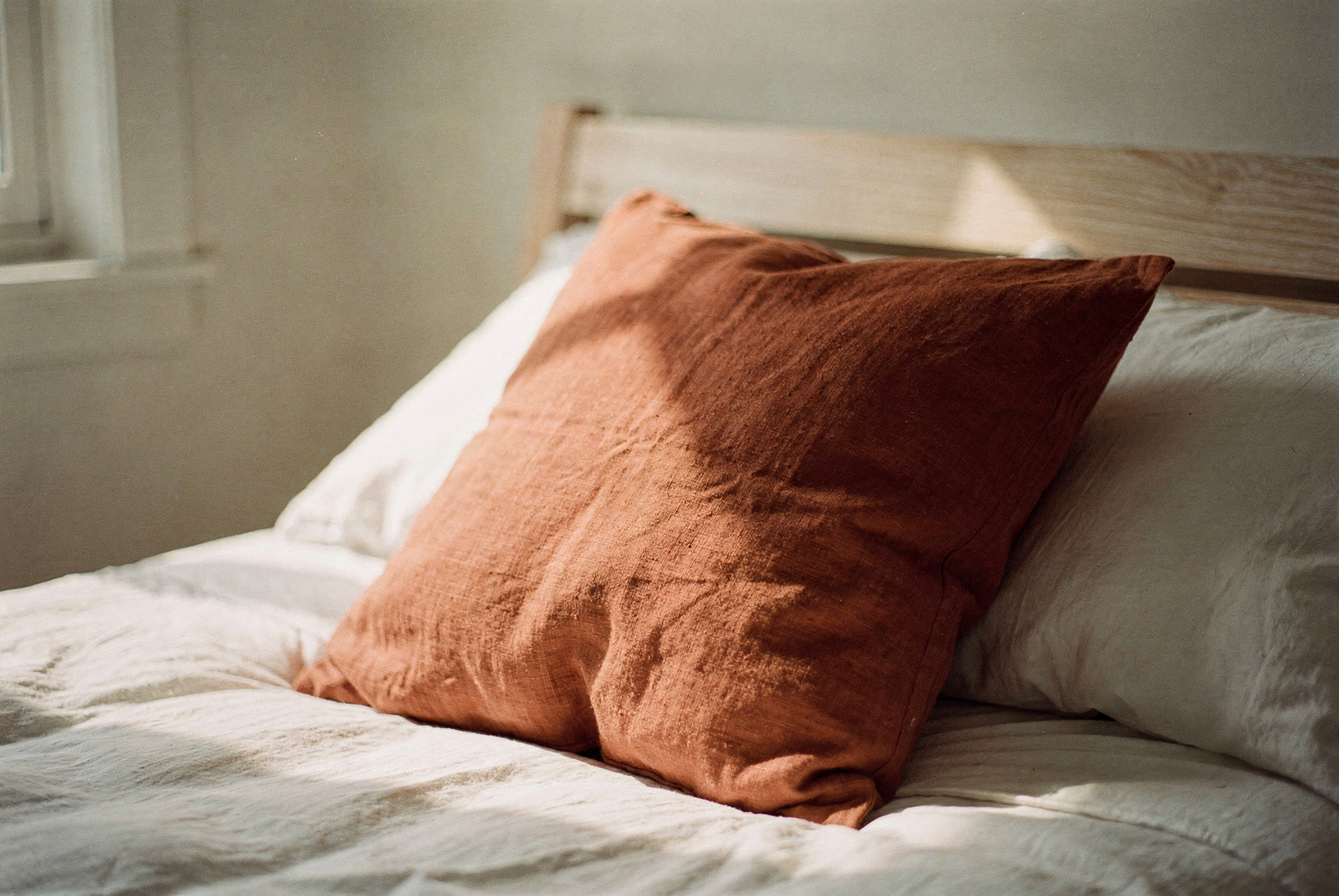 A single textured terracotta throw pillow on crisp white linen bedding, casually angled, natural morning light, simple wooden bed frame, editorial interior photography