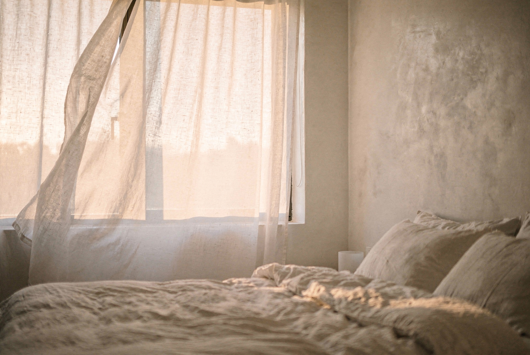 Sheer white linen curtains billowing gently in a summer breeze, large bedroom window, soft filtered morning light, minimal white bedding, airy and calm atmosphere, editorial interior photography