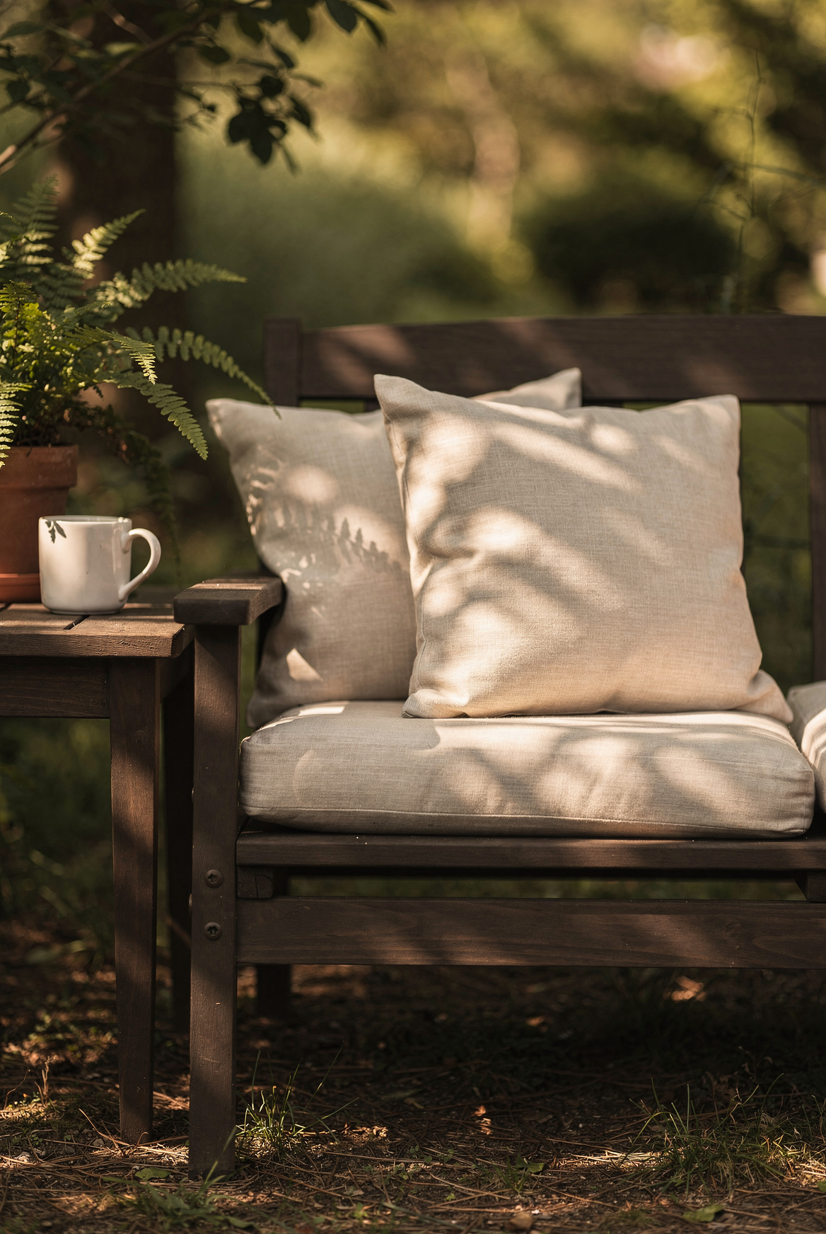Two thick beige outdoor cushions on a dark wood bench, a small side table with a ceramic mug and a fern, soft afternoon light filtering through tree leaves, cozy garden seating area