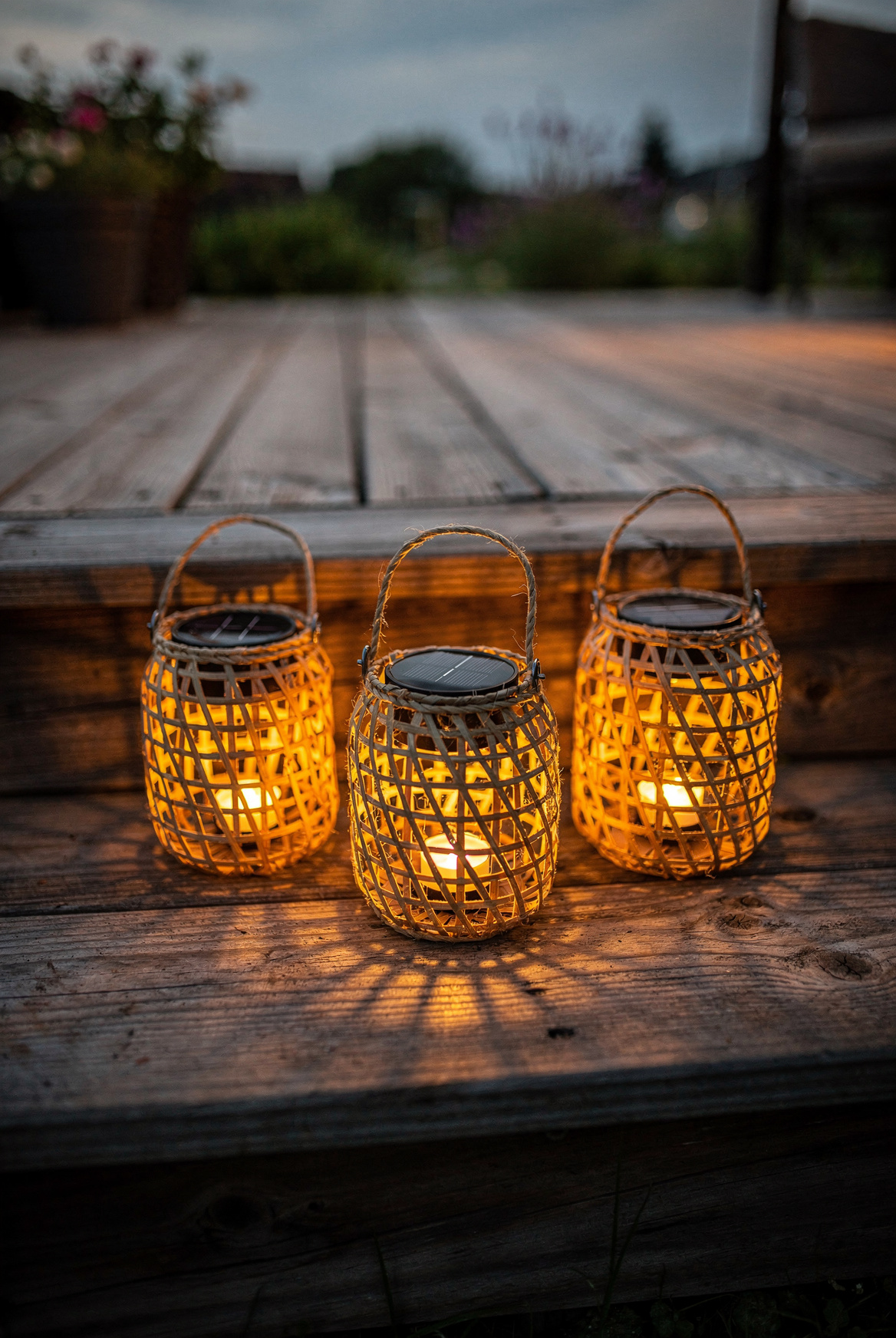 Three small woven solar lanterns clustered on wooden patio steps, warm golden glow inside each, dusk light, soft amber illumination, cozy summer evening on a deck