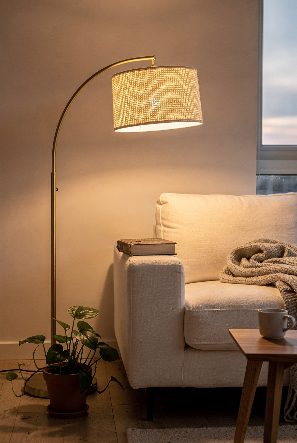 A Brightech Logan gold arc floor lamp with woven drum shade arcing over a cream sofa in an apartment corner, warm amber glow, overhead light off, pothos plant at the base