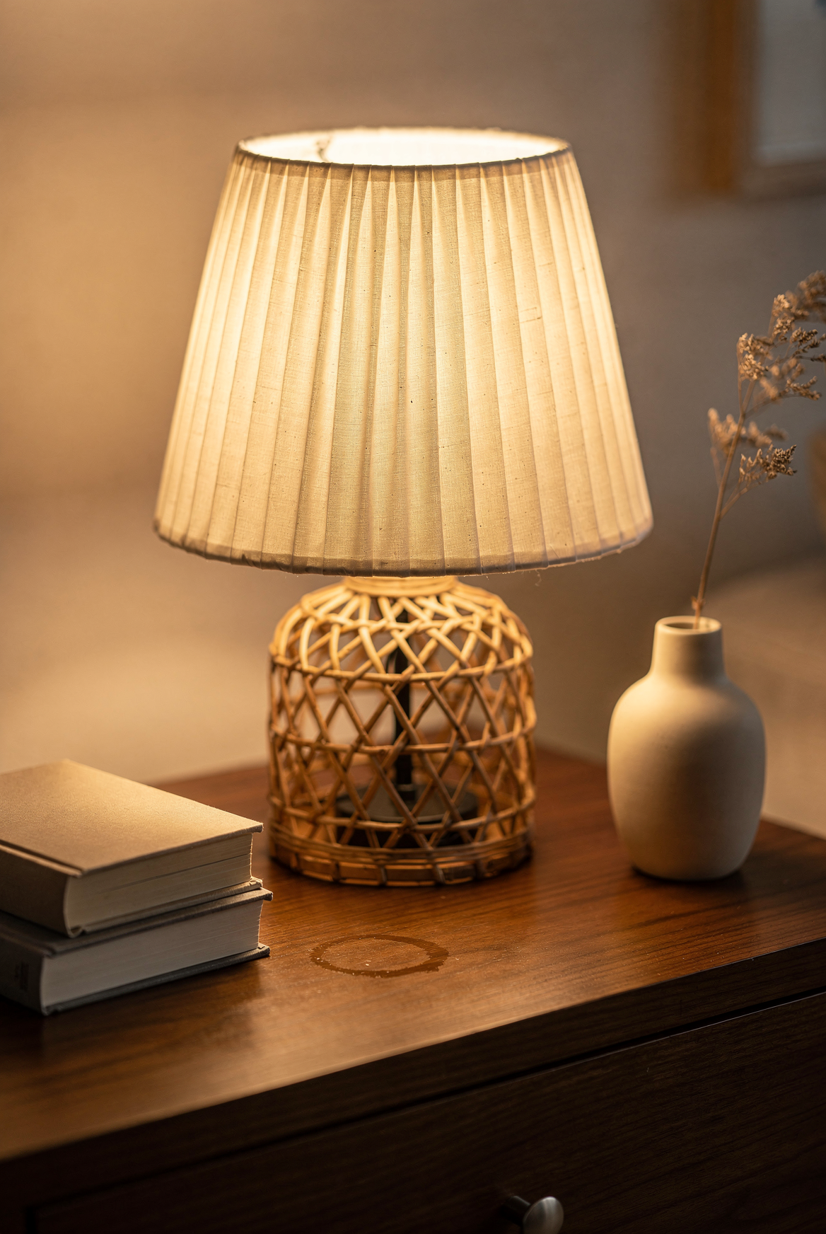 Close-up of a kvppvt pleated cream table lamp with rattan base glowing warm amber on a wood side table, stack of books, ceramic bud vase with dried stem alongside